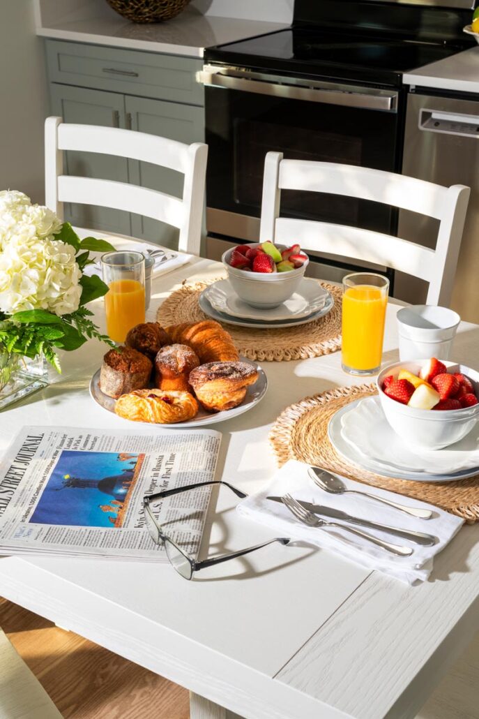 Kitchen dining table in the sunlight with muffins, orange juice, bowl of fruit and a newspaper next to reading glasses