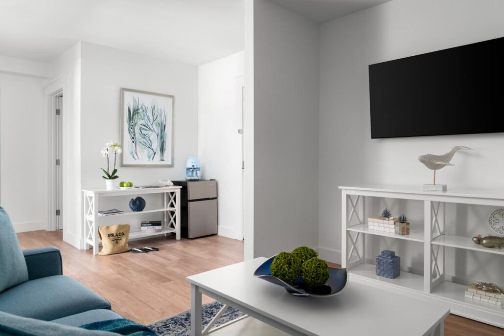 Living room with sofa, coffee table, tv on the wall with stand underneath and a entryway with bench and a small refrigerator