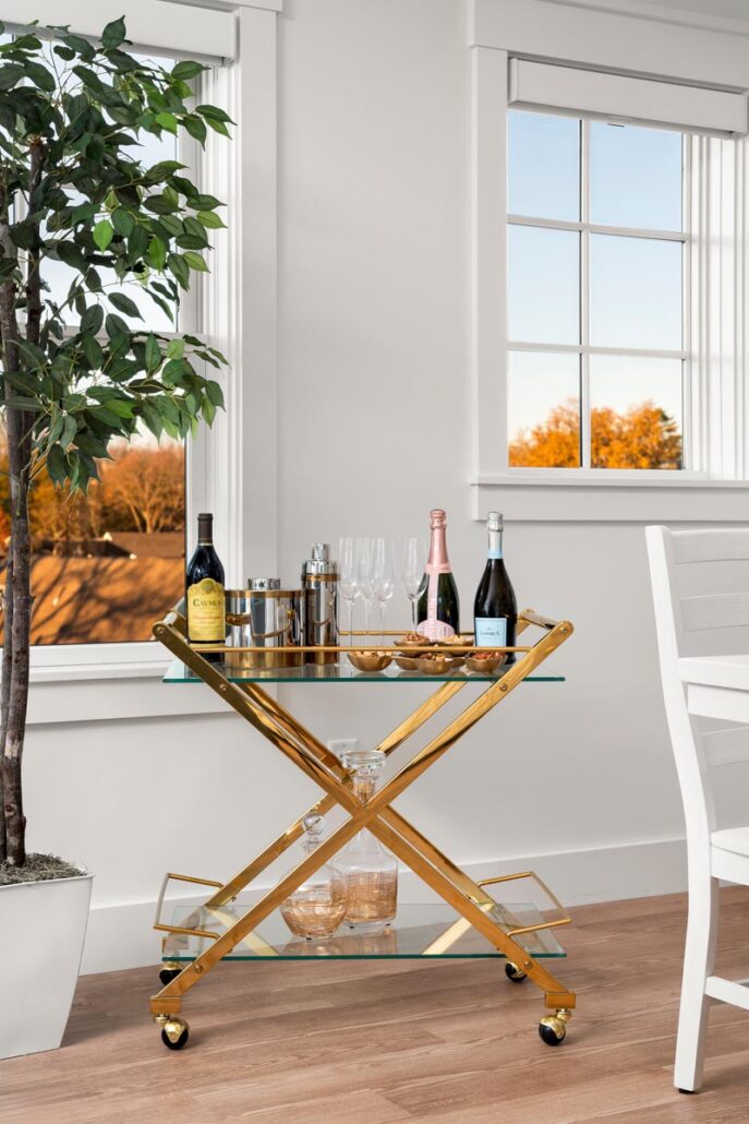 Living room with a bar cart that has bottles of alcohol and drinking glasses on top next to a large plant in front of two windows overlooking trees