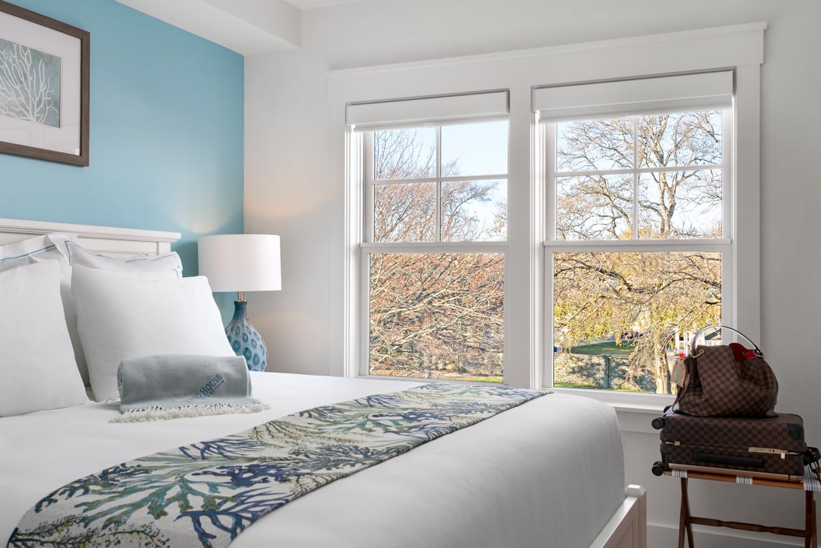 Room with bed dressed with white linens and a Louis Vutton luggage and purse next to it facing a window overlooking trees