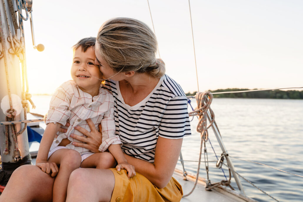 Mother kisses her smiling son on the cheek while sitting on a boat, with the sun setting over the water in the background.