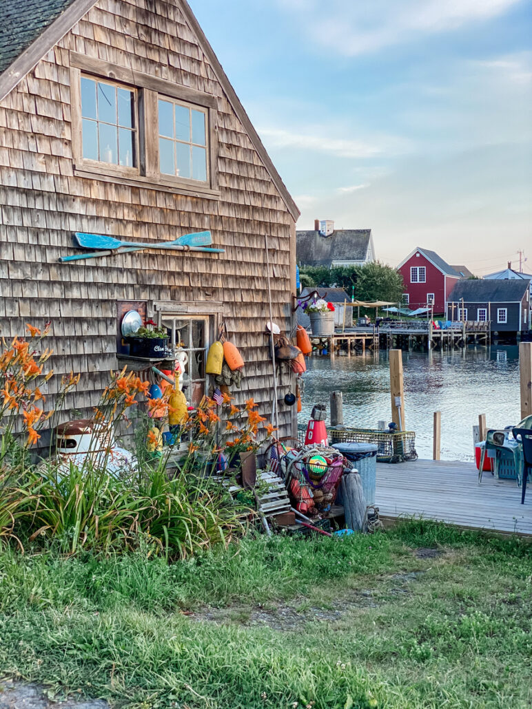 "Rustic shingled building by the water, decorated with colorful buoys and fishing gear, surrounded by greenery and flowers near a dock.