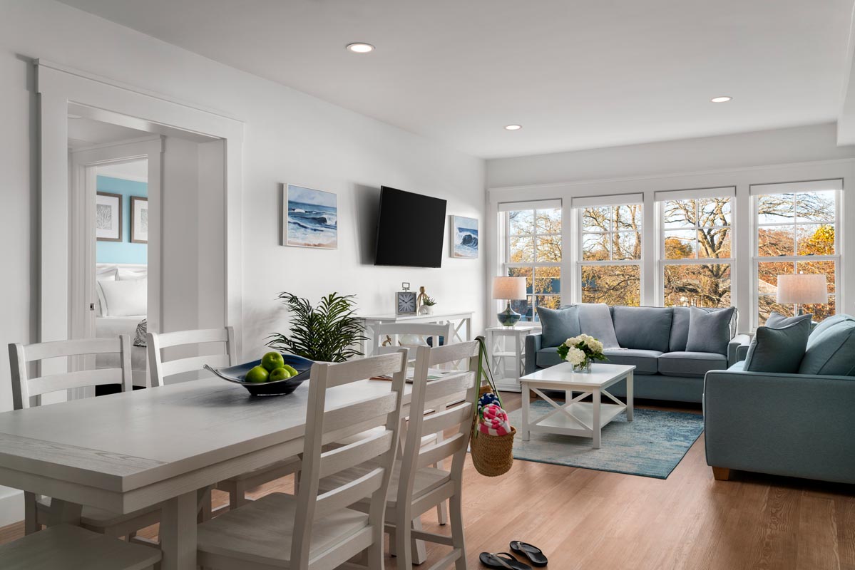 Spacious living room with comfortable sofas, a TV, and a dining table, featuring a large window with a view of lush trees.